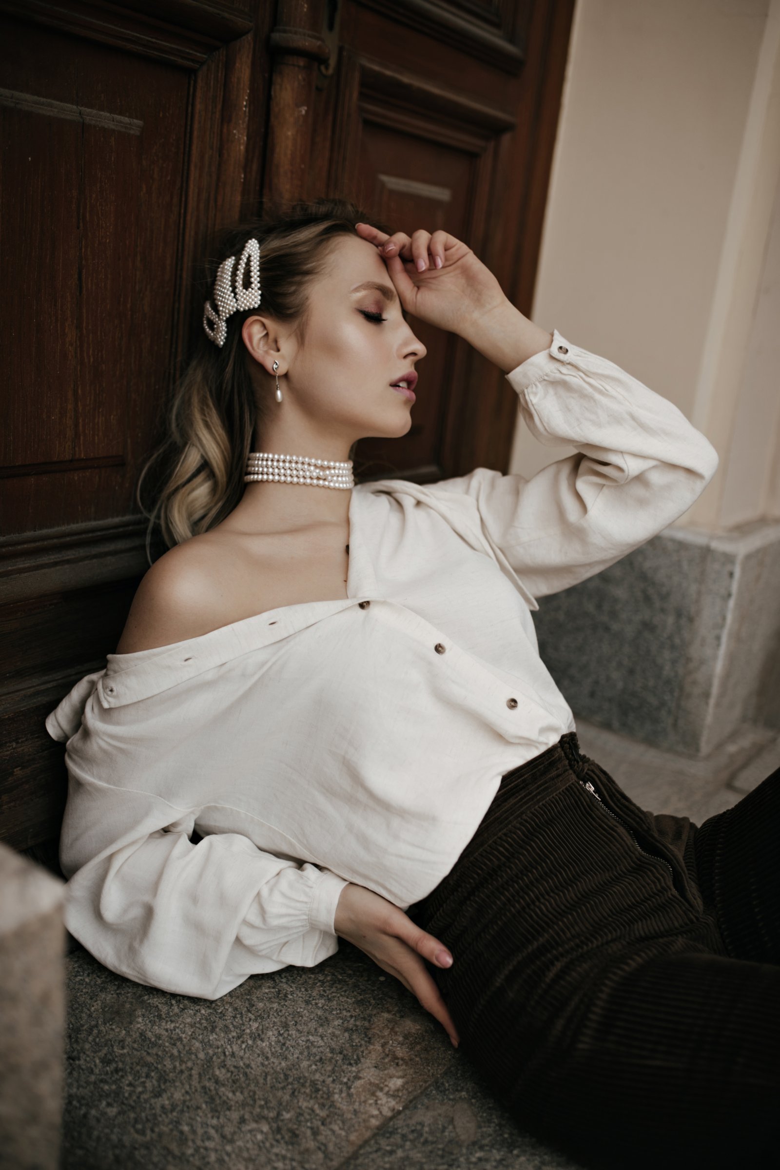 Charming curly blonde woman in pearl jewelry, white blouse and brown velvet pants sits near wooden